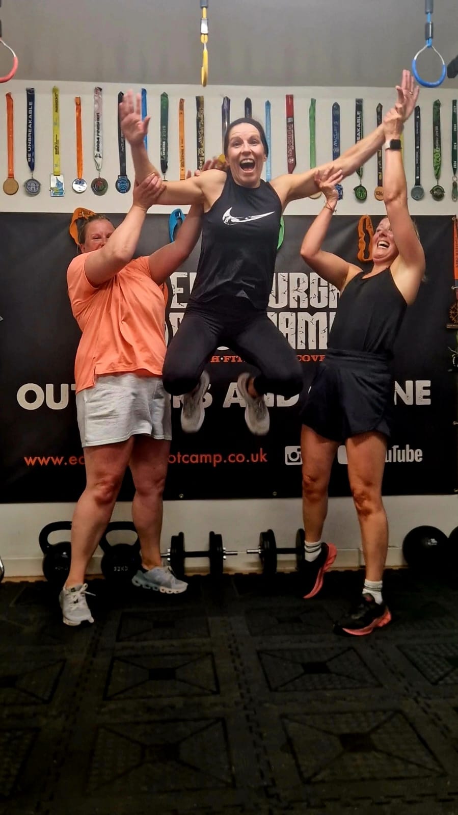 Members mid-jump at the EBC Gym in front of a wall of race medals, kettlebells and dumbbells racked behind