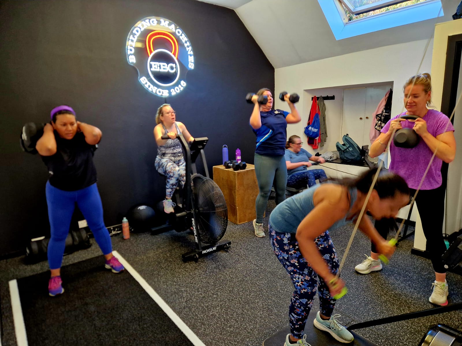 A small-group session in the EBC Gym: sandbag, air bike, dumbbells, rower and kettlebell under the neon EBC sign